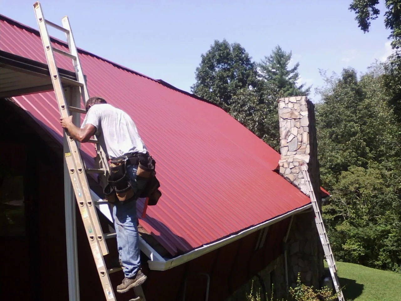 Roofer on a ladder beside a red metal roof and stone chimney — Soffit & Fascia Repair by Roofing Brothers in Atwood